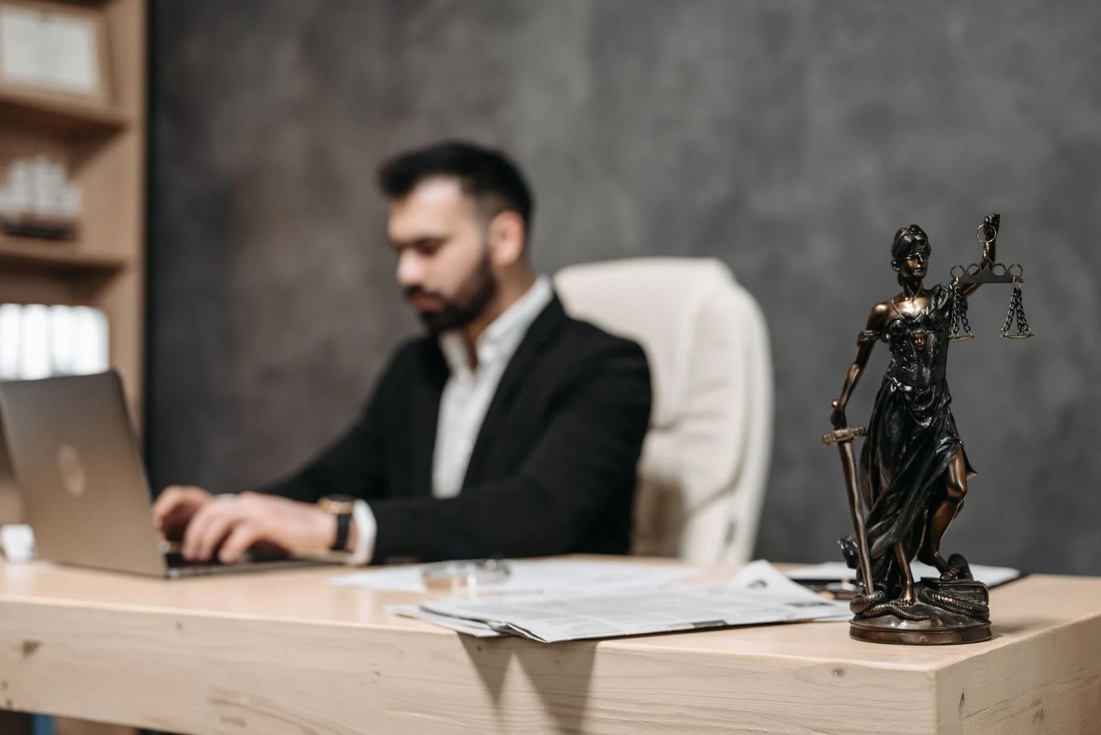 A businessman works on a laptop at a desk, with a Lady Justice statue in focus beside him.
