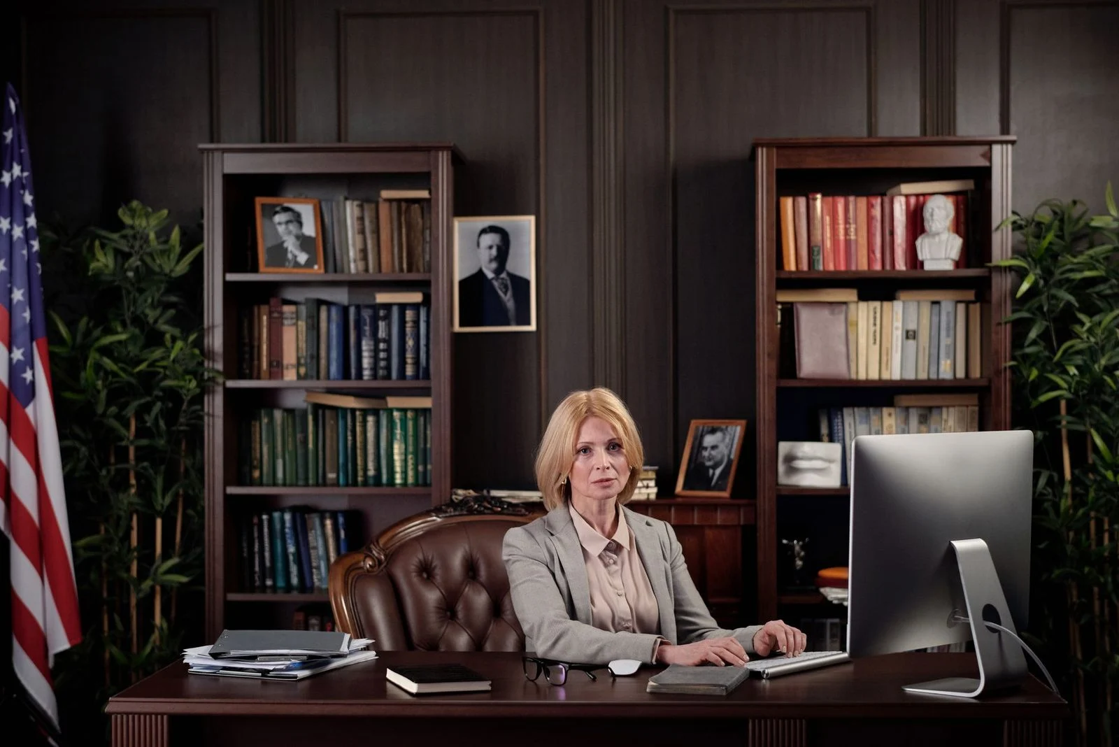 A businesswoman sits confidently at a desk in an office with bookshelves and an American flag.
