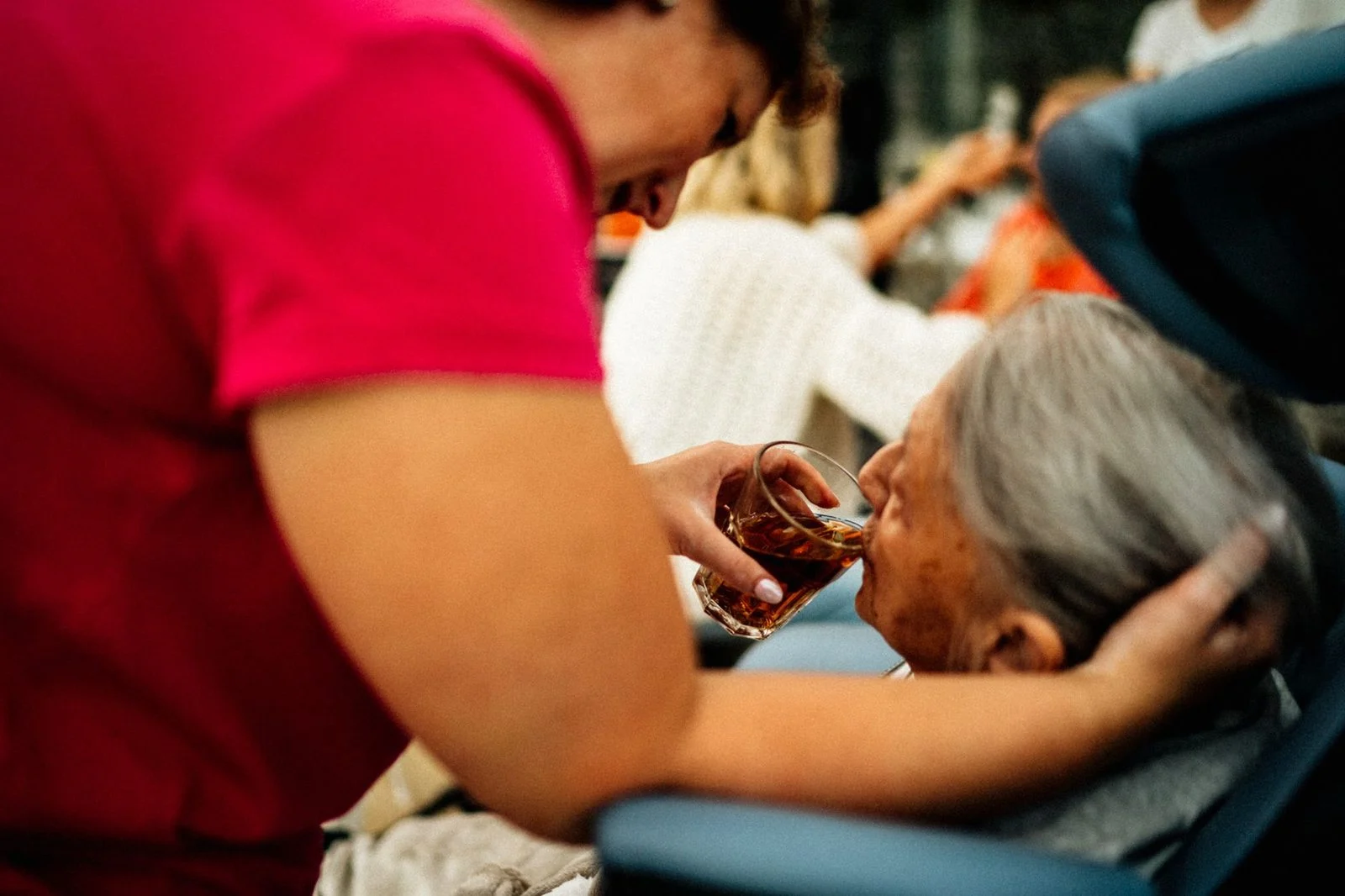 A caregiver helps an elderly woman hold a cup of tea at a kitchen table.