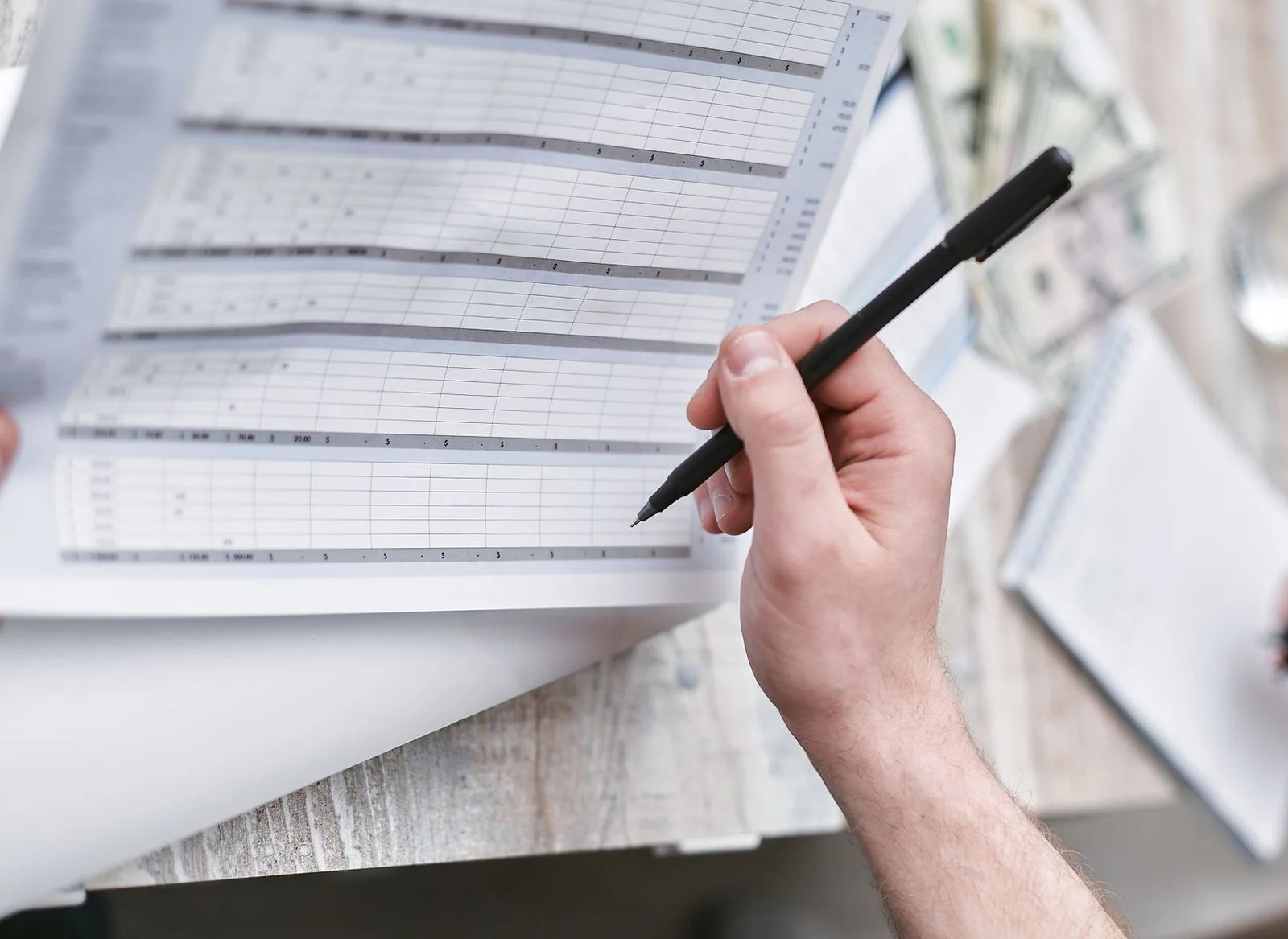 A close-up of a hand holding a pen over a financial document with cash on the desk.