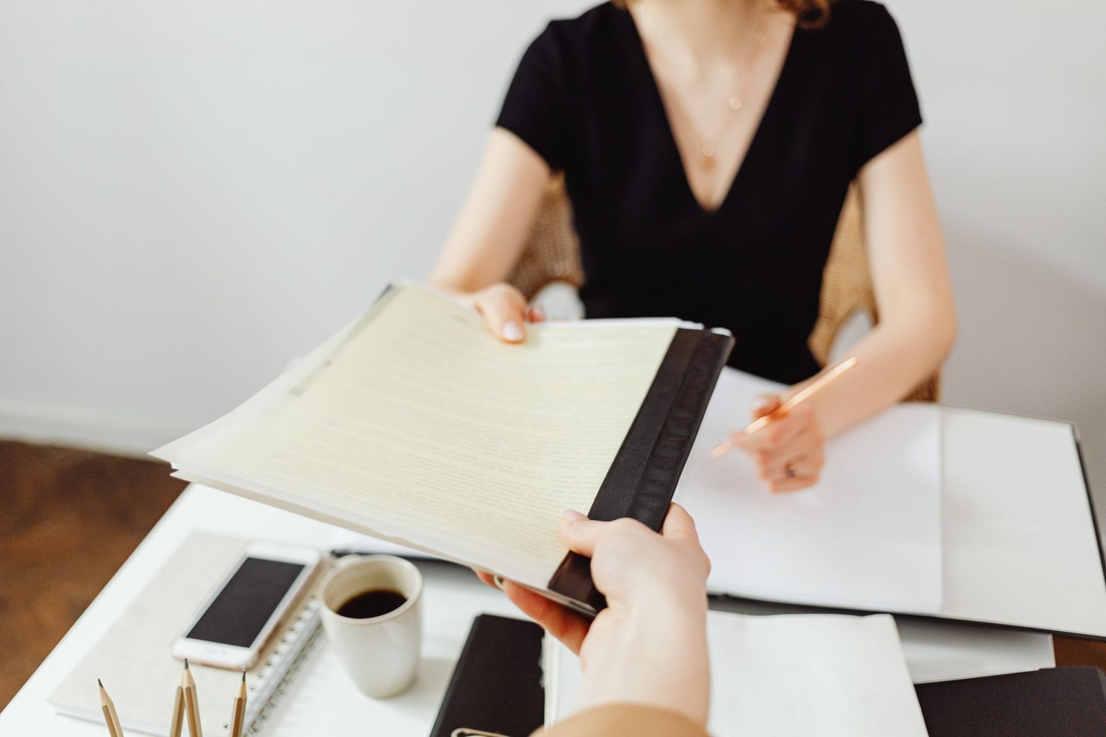 A close-up of hands passing a printed document across a desk beside a pen and laptop.
