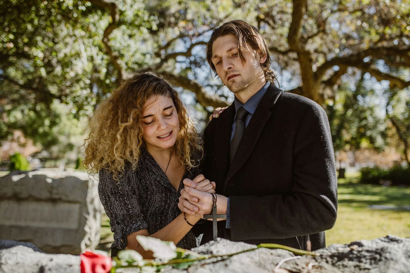 A couple holds hands while standing together at a grave in a quiet, peaceful cemetery.