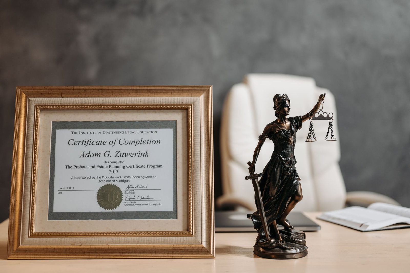 A framed legal certificate and a Lady Justice figurine sit on a wooden desk in a law office.