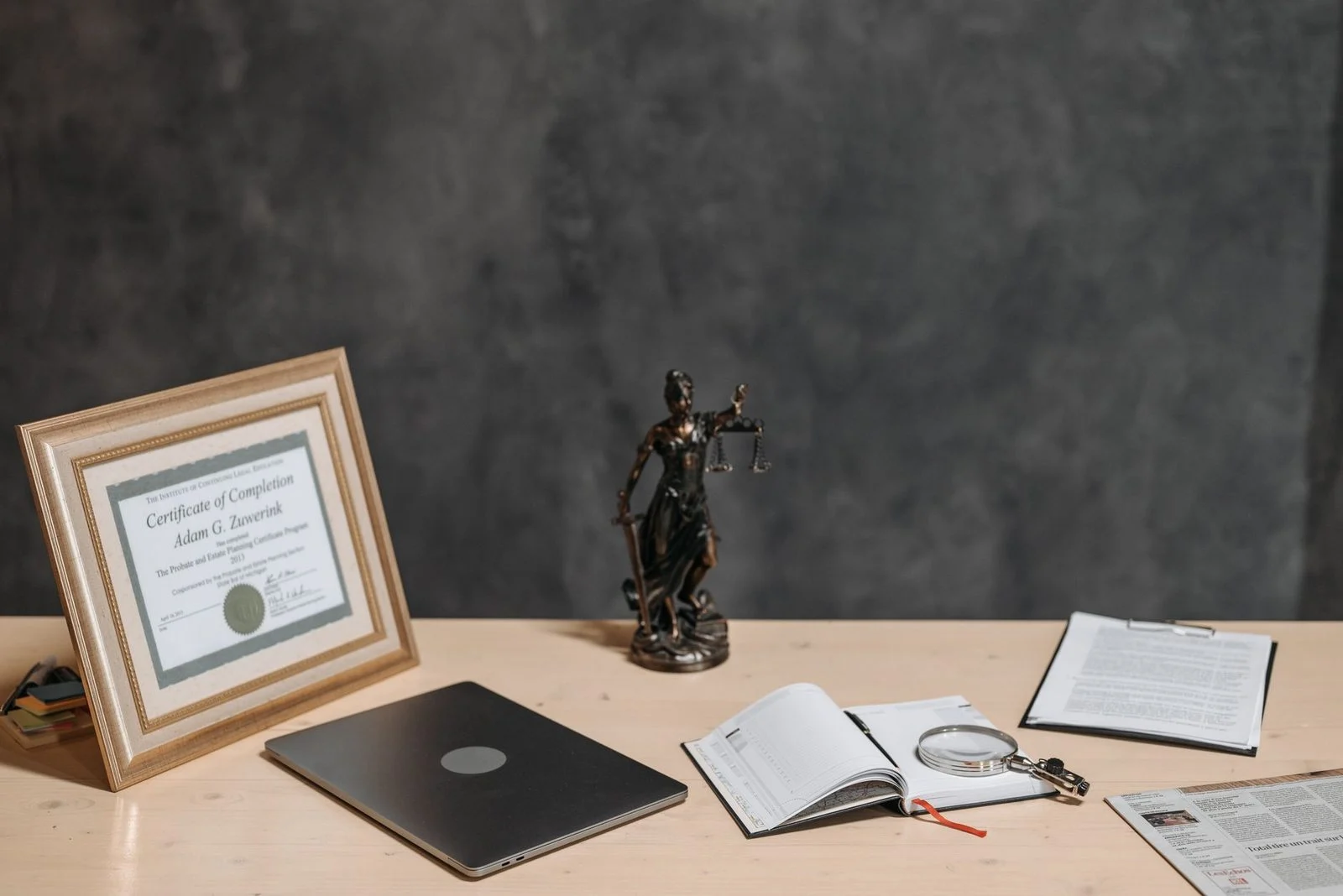 A lawyer's desk with a justice statue, diploma, laptop, magnifying glass, and documents.