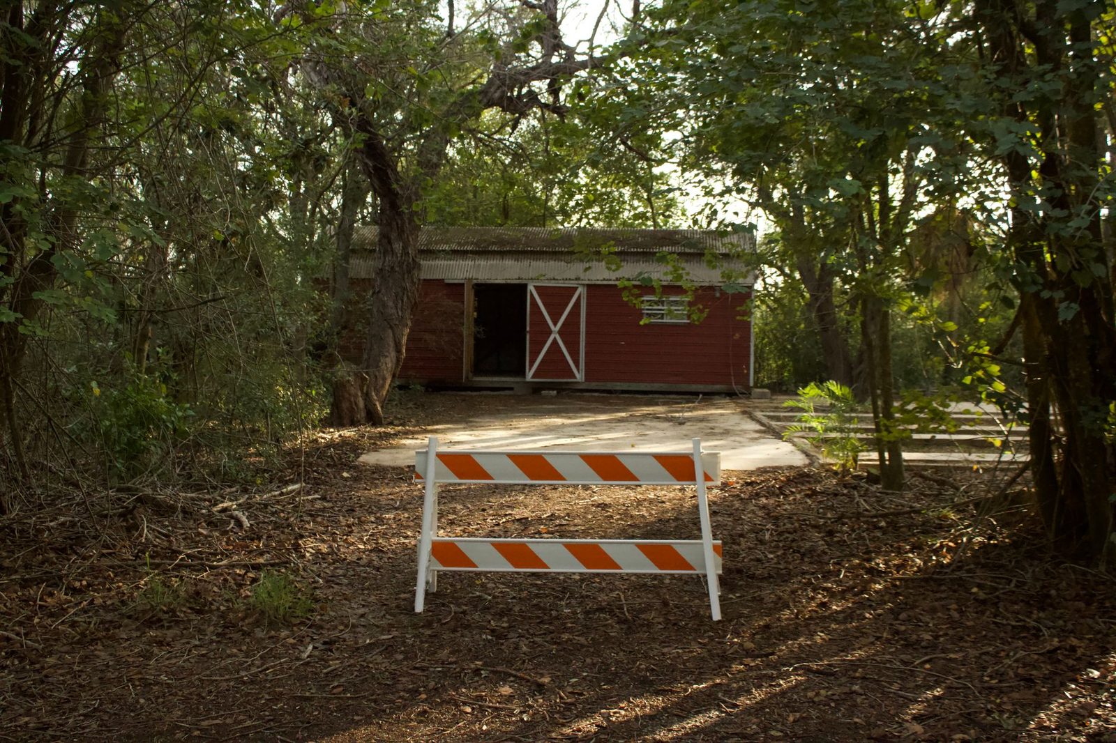 A secluded red barn stands deep in dense forest, with a no-entry barrier blocking the path leading to it.