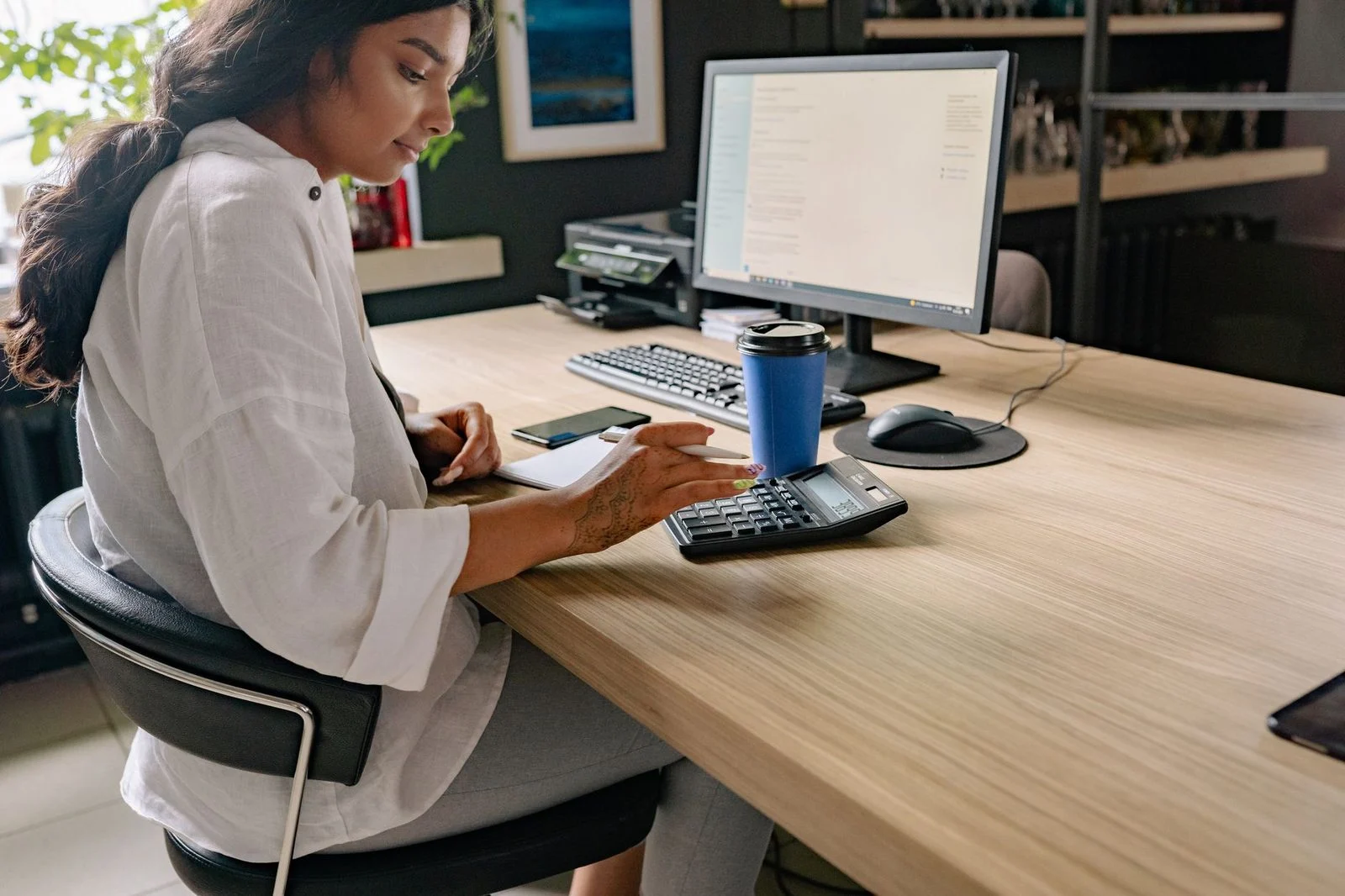 A woman sits focused at an office desk, working on a calculator and computer.