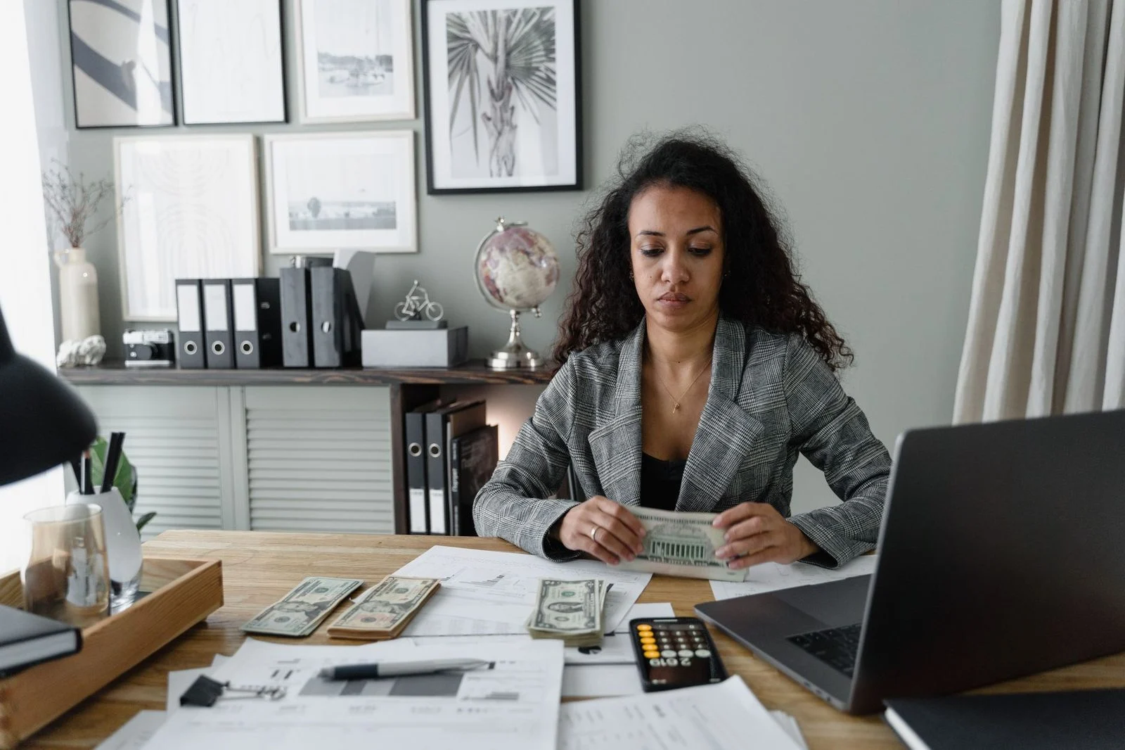 A woman sits at a desk counting cash beside office supplies.