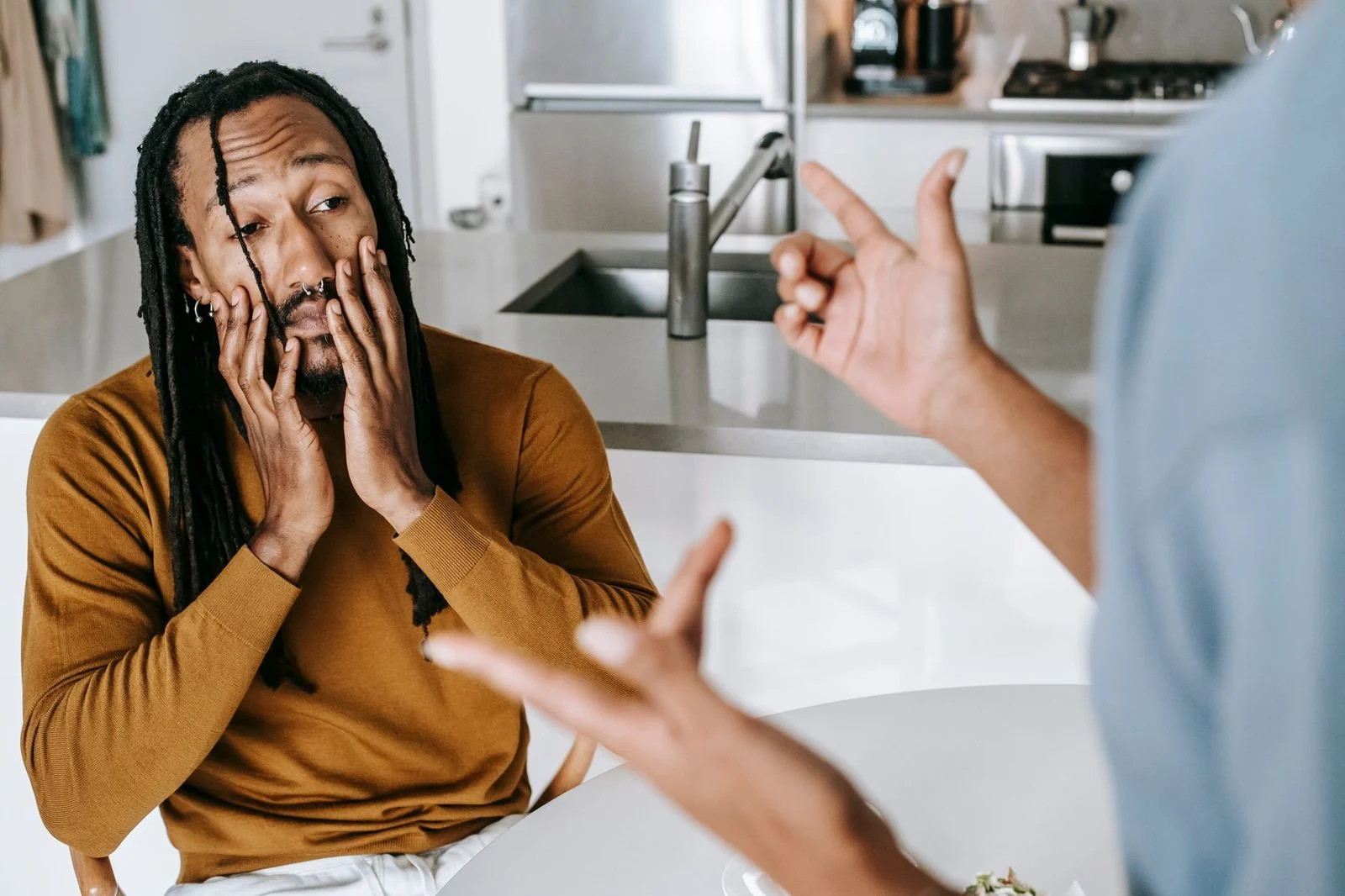 A young African American man sits at a kitchen table with his hands on his face while a woman stands nearby in apparent conflict.