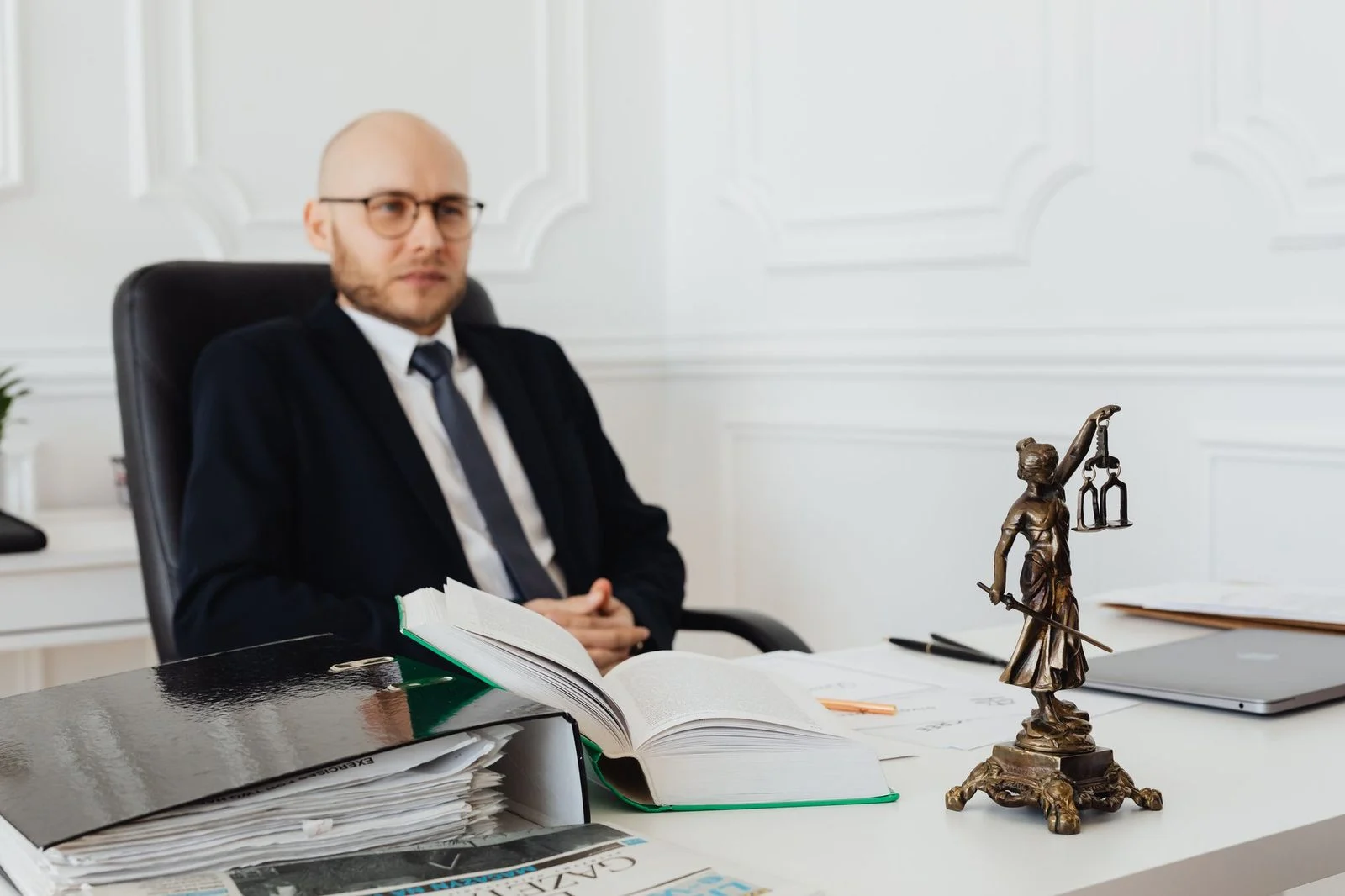 A bald lawyer sits in an office with legal documents beside a statue of Lady Justice.
