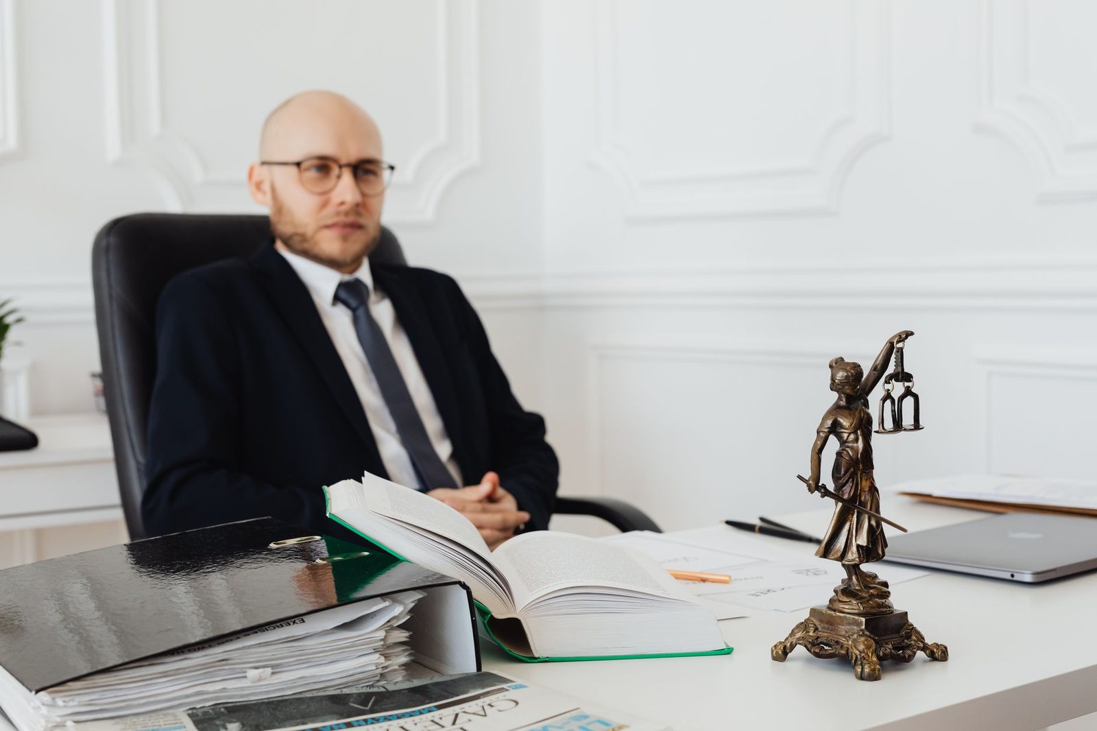 Bald lawyer seated in an office with legal documents beside a Justice statue.