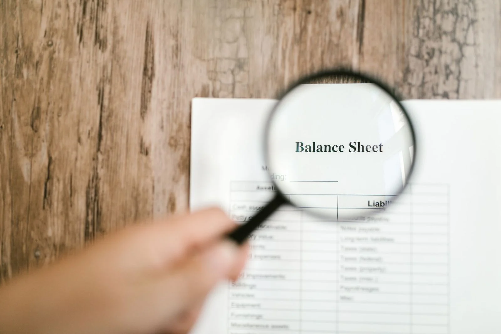 Close-up of a hand holding a magnifying glass over a financial balance sheet on a wooden desk.