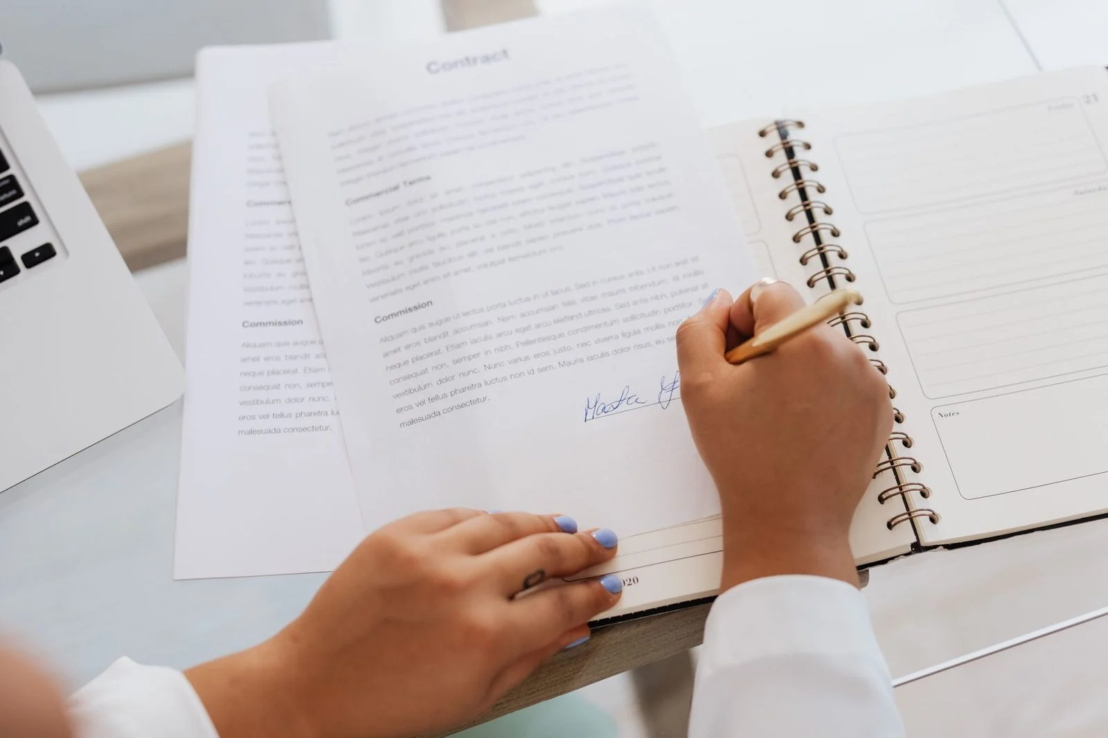 Close-up of a person's hands signing a document on a desk beside pens and office supplies.