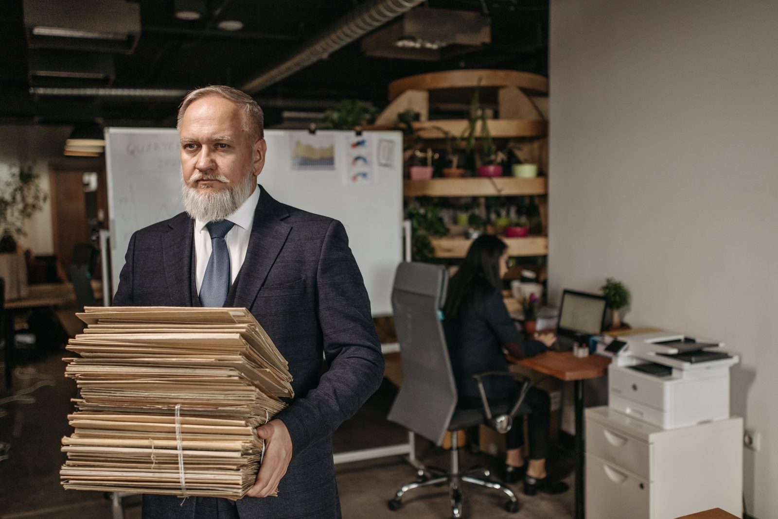 Elderly man in a suit holding a large stack of documents in a modern office.