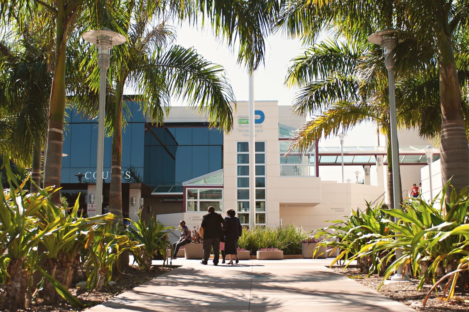 Exterior of a Miami courthouse with palm trees and people walking under a bright blue sky.