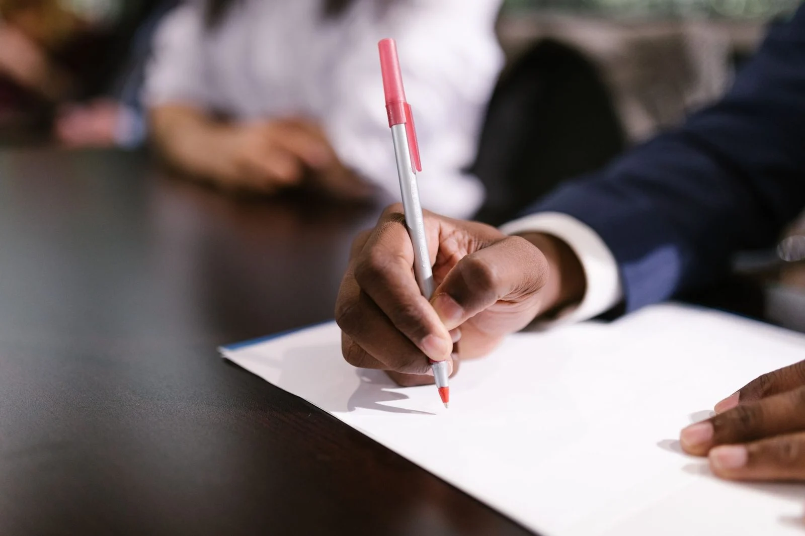 Hand holding a pen while writing on white paper at a desk.