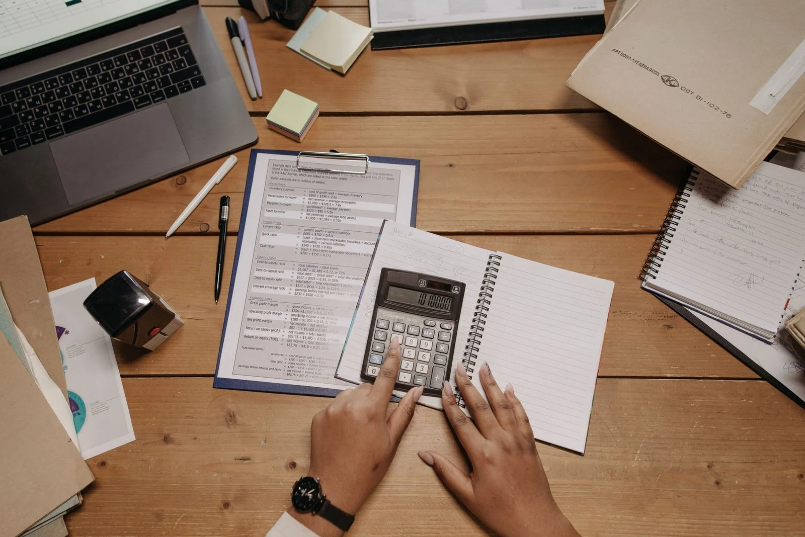 Hands using a calculator beside stacked documents on a wooden desk.