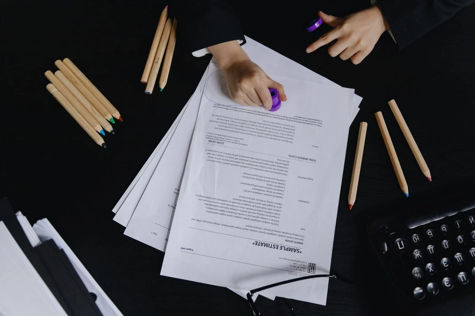 Hands writing with colored pencils on documents on a black desk.