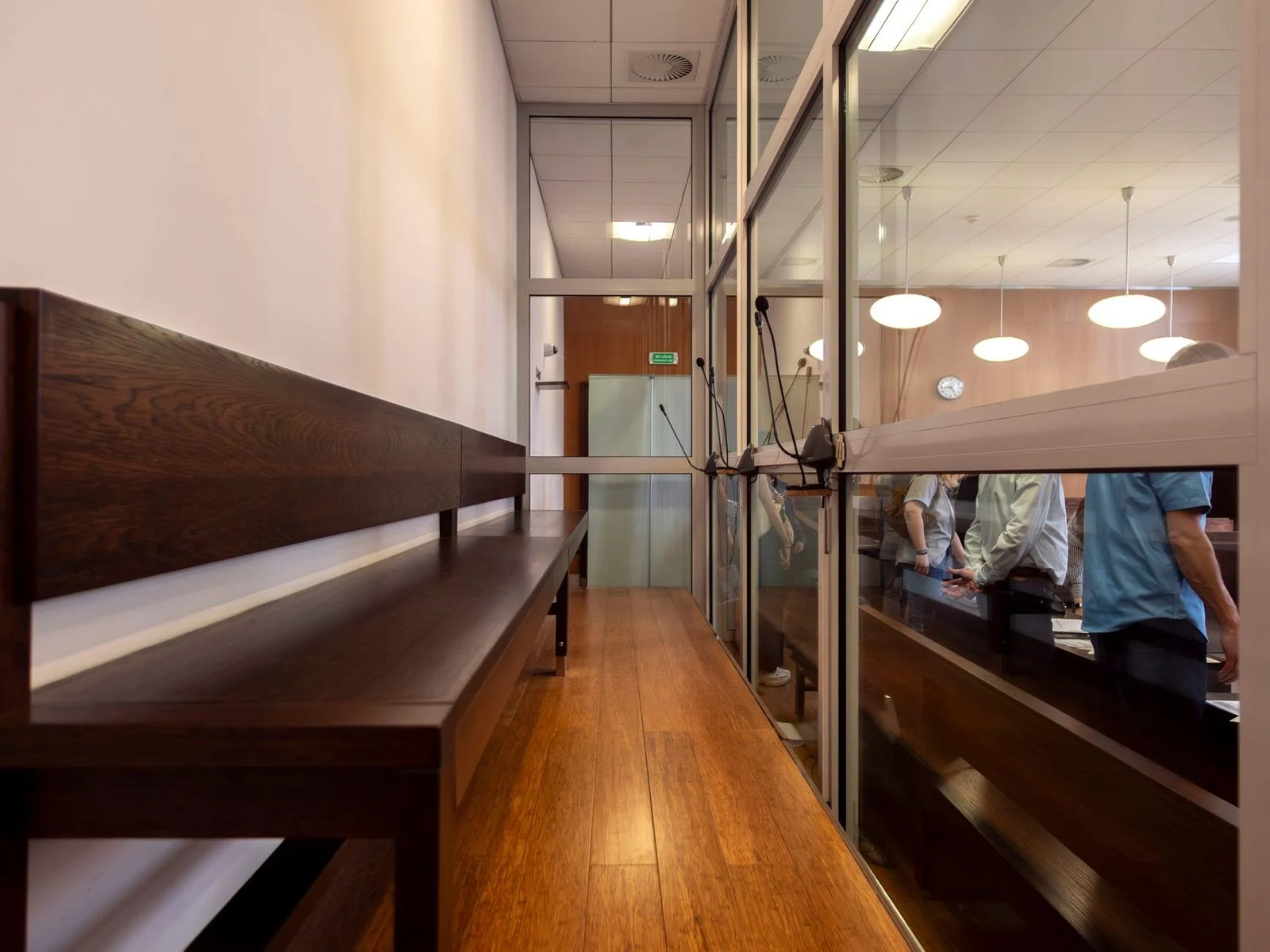 Empty courtroom with wooden benches and glass partitions, viewed from the public gallery.