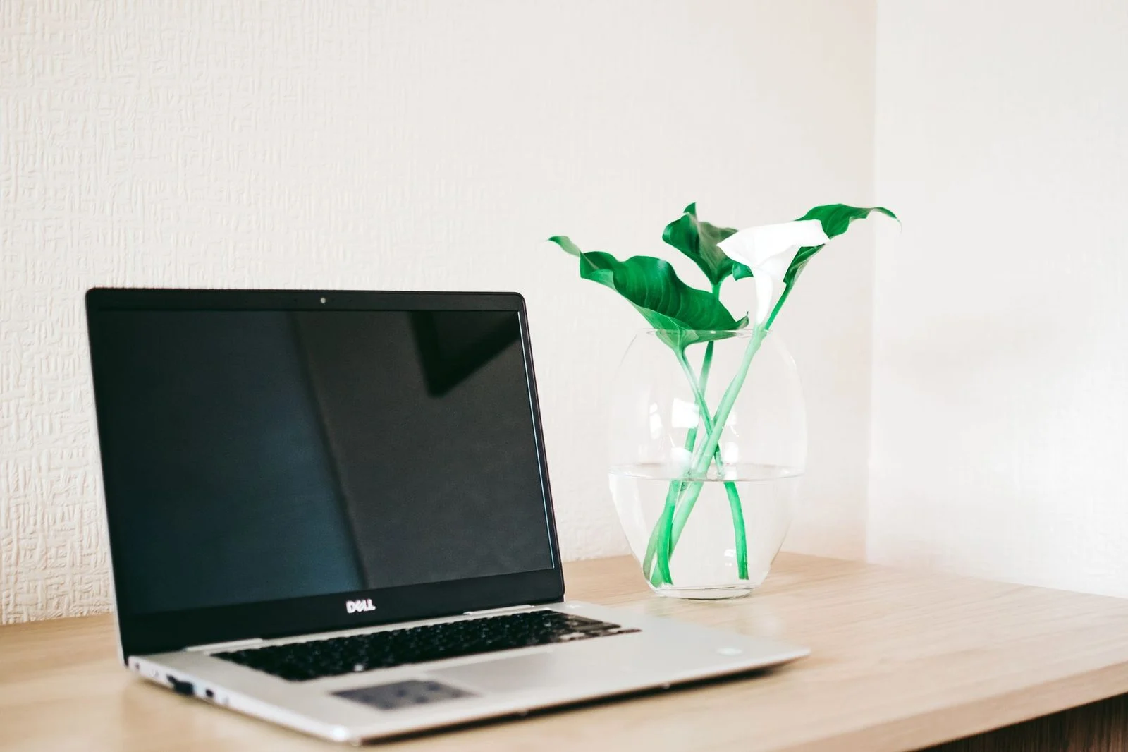 Laptop on a wooden desk beside a vase of green and white flowers.