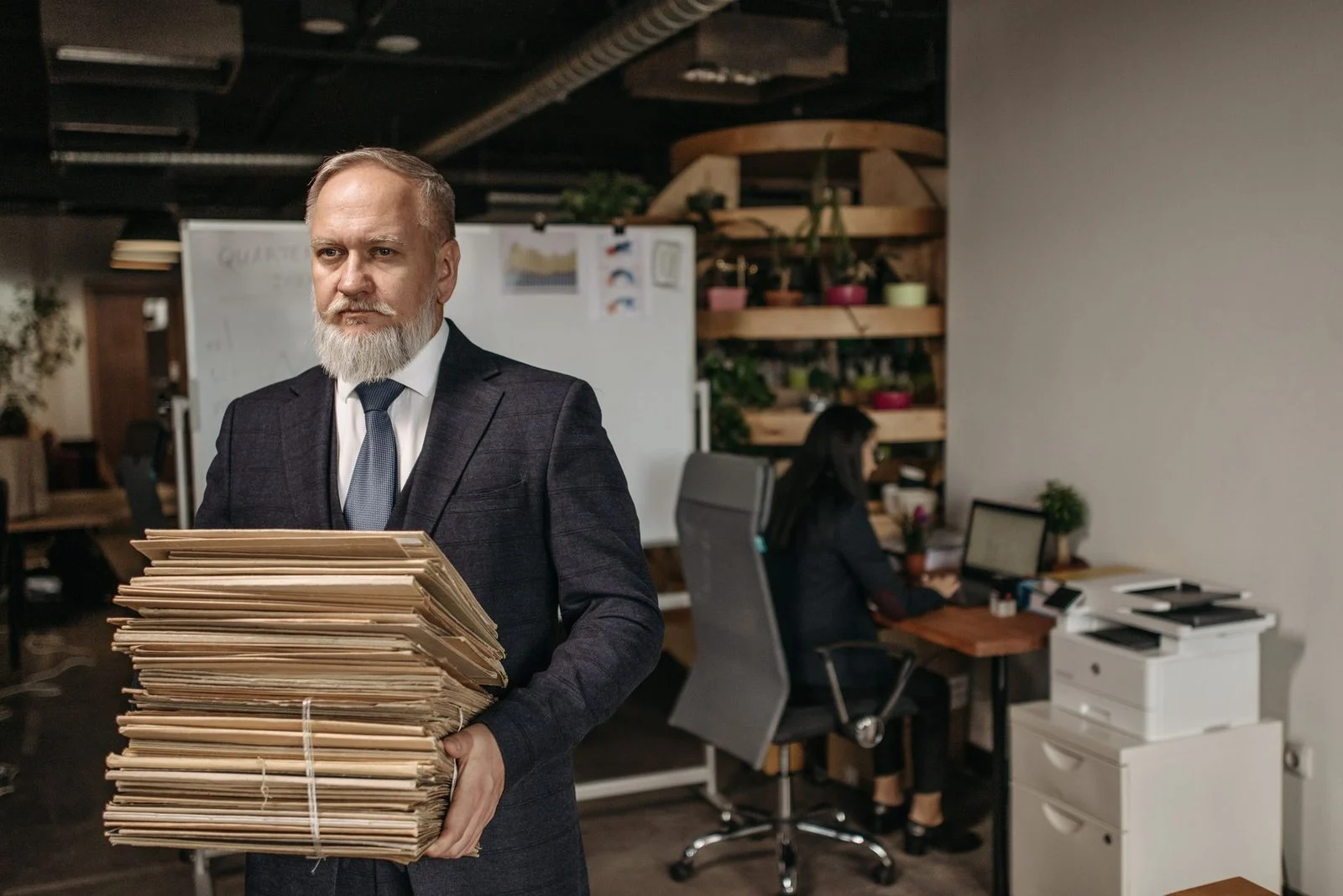 An older man in a suit holds a large stack of documents in a modern office.