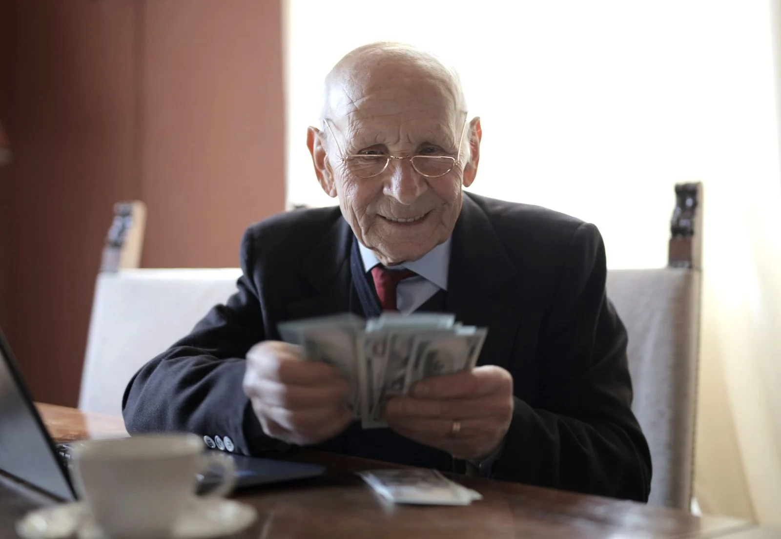 Smiling senior man in a suit and eyeglasses counts cash at a wooden table beside a laptop and a cup.
