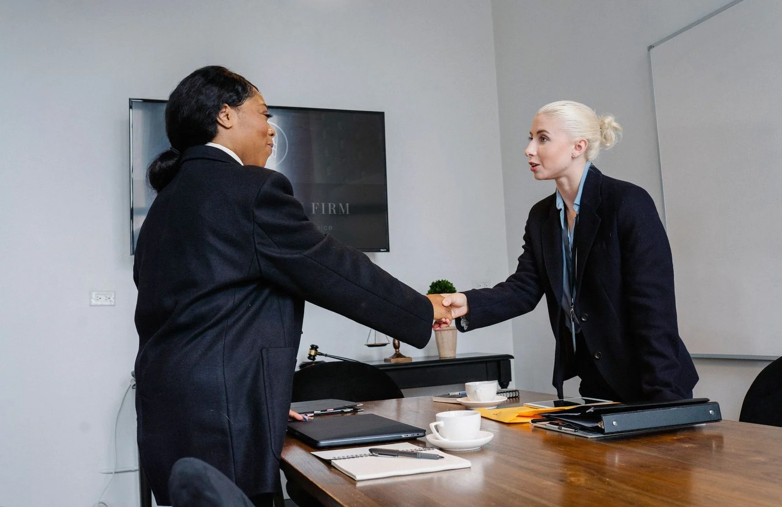 Smiling women in formal wear shake hands over documents on a table.