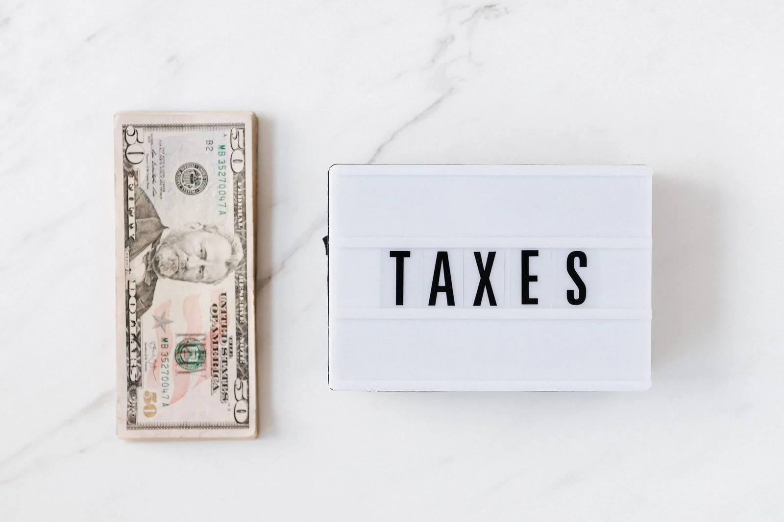 Stacked US dollar bills beside a glowing white light box reading "TAXES" on a white marble surface.
