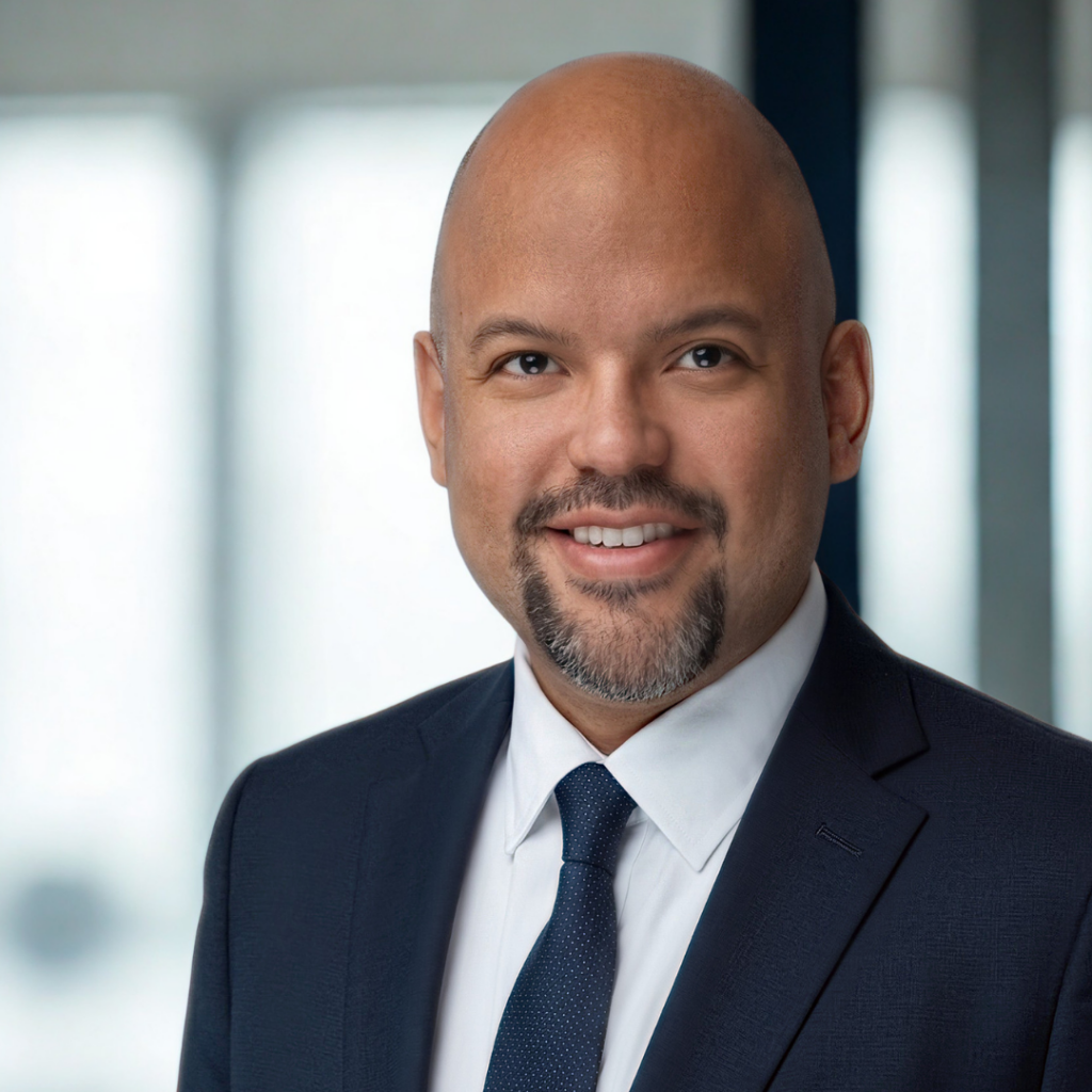 Professional headshot of a smiling bald man in a navy suit and white shirt, wearing a dark tie, in a bright office setting.