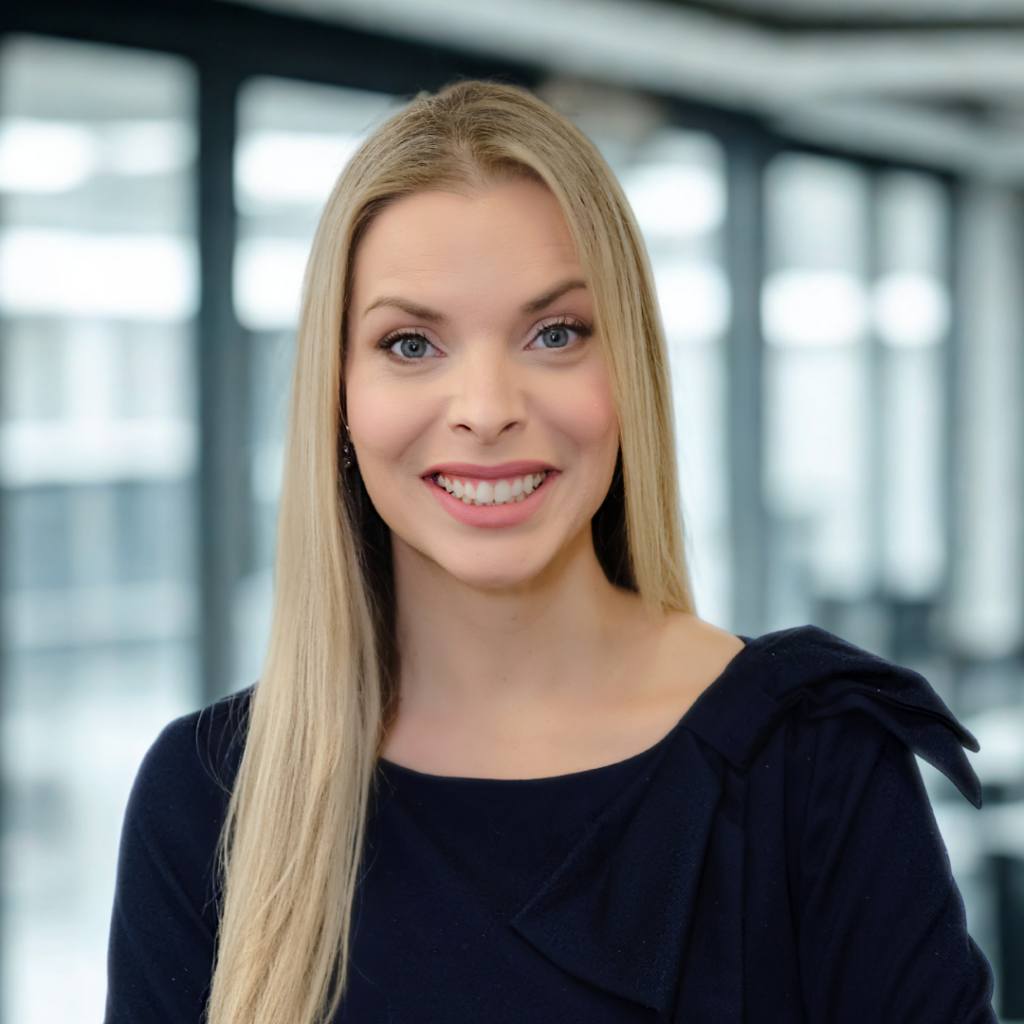 Smiling blonde woman in a navy blouse posing for a professional portrait in a bright office setting.