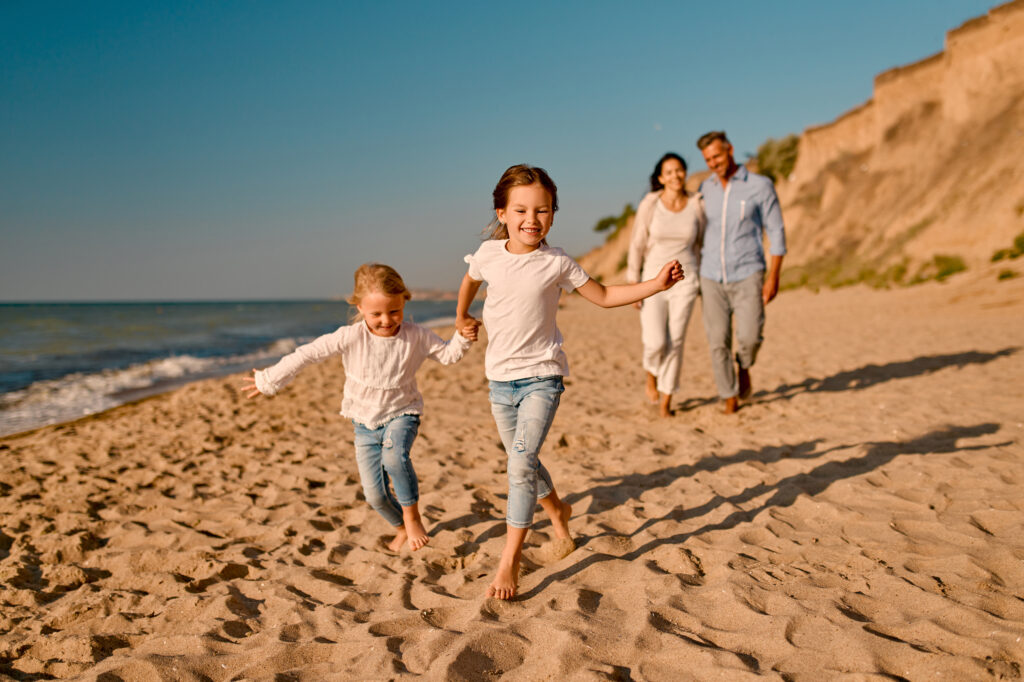 Two young girls run barefoot along a sandy beach while their smiling parents walk behind, blue sky and rocky cliffs in the background.