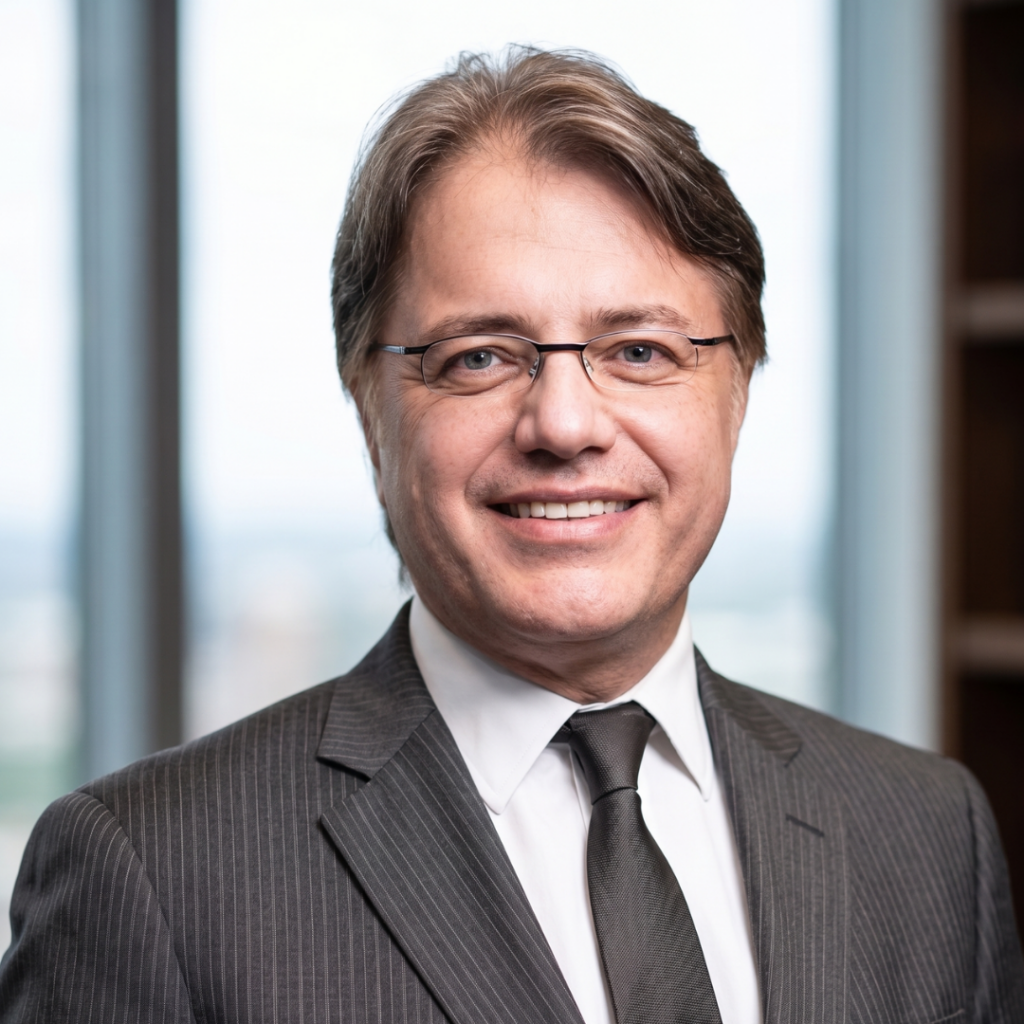 Smiling professional headshot: man in a gray pinstripe suit, white shirt, black tie, wearing glasses in an office setting.